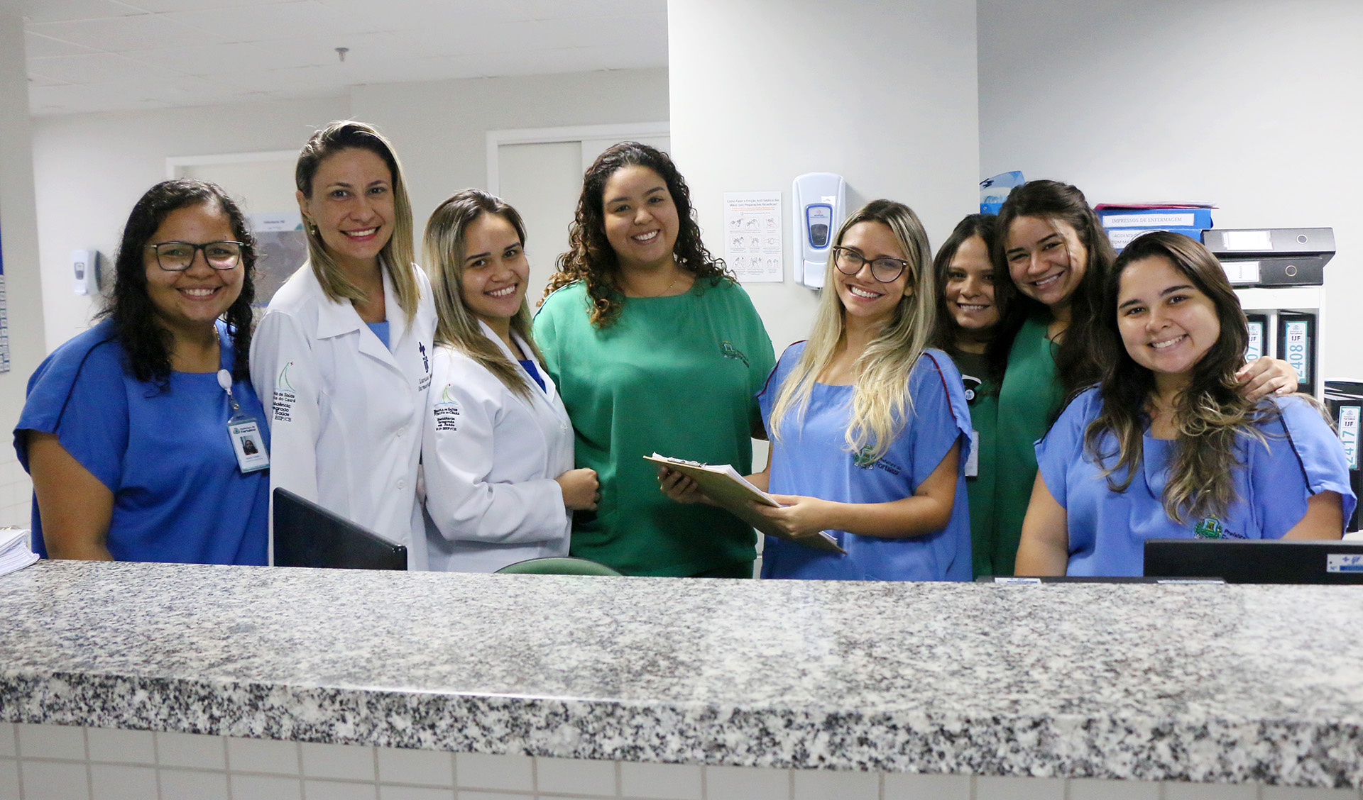 moças de jaleco e batas sorrindo para a foto atrás de balcão de hospital